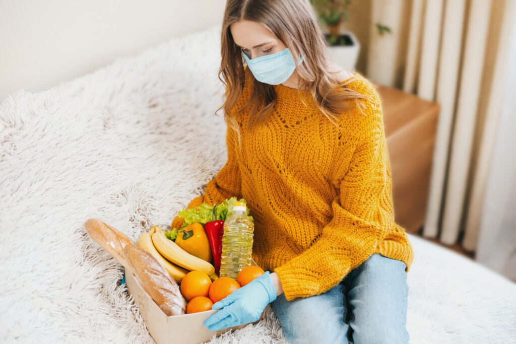 young girl volunteer medical mask holds cardboard box with vegan food fruits vegetables sits couch home delivery coronovirus quarantine stay home concept